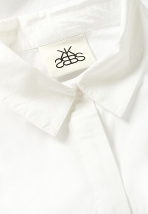 Bela bombažna srajca s koničastim ovratnikom, sprednjim gumbnim delom in tkaninsko oznako z črnim logotipom, ušito znotraj ovratnika.