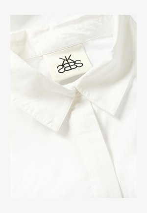 Valge puuvilla särk terava kraega, eesmise plaaniga ja kangasildiga, millel on must logo õmmeldud krae sisse.