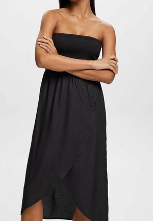 Nainen, joka pukeutunut olkaimettomaan mustaan midimekkoon, jossa on kerätty yläosa ja päällekkäinen tulppaanikuvioitu hame, kädet ristissä rinnan edessä.