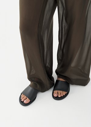 Lábak fekete papucsban, bő, áttetsző barna nadrággal, sima fehér háttér előtt.