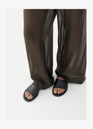 Pėdos juodose laisvų sandalų poroje, dėvint laisvus, permatomus rudus kelnes, fone paprasta balta spalva.