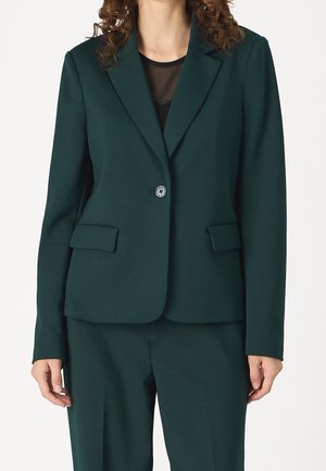 Kvinde iført en mørkegrøn blazer med enkelt knap og laplommer samt matchende bukser over en sort gennemsigtig top.
