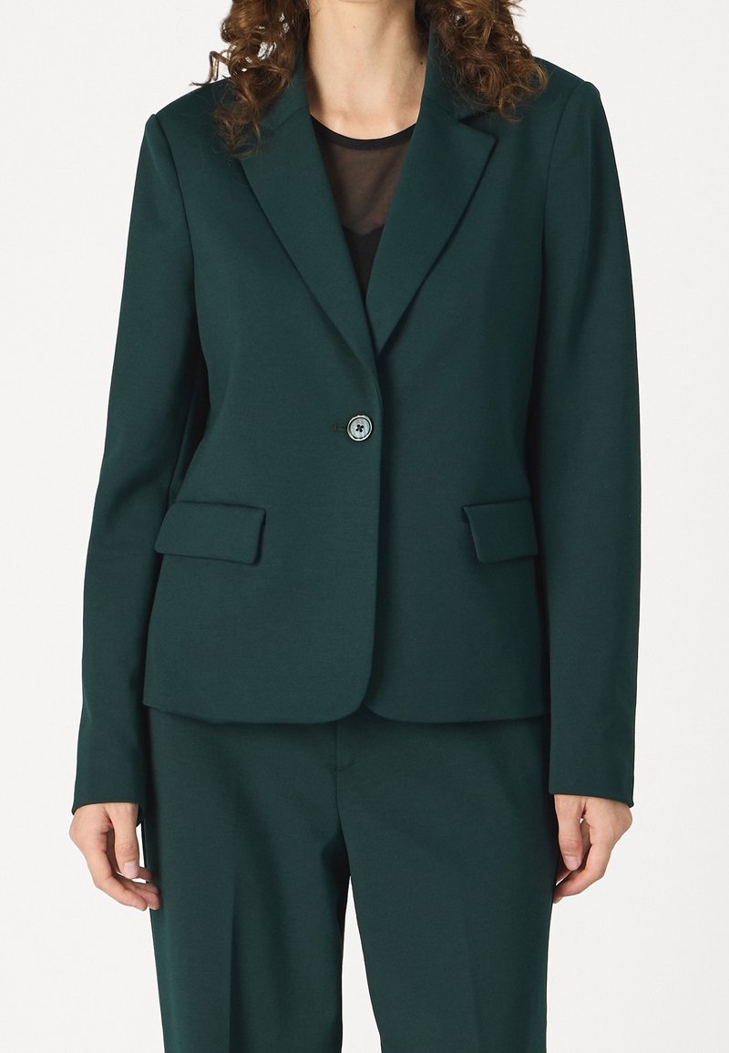 Femme portant un blazer vert foncé à un bouton avec poches à rabat et pantalon assorti sur un top noir transparent.