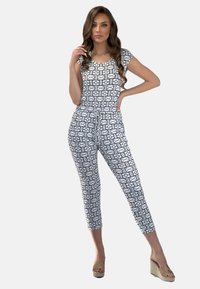 Biały kombinezon z geometrycznym czarnym wzorem, z krótkimi rękawami, okrągłym dekoltem i zwężanymi, skróconymi nogawkami, zestawiony z beżowymi sandałami na platformie.