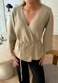 Beige Wickelstrickjacke mit 3/4-Ärmeln, V-Ausschnitt, Kordelzug in der Taille, aus weichem Stoff, mit gerippten Akzenten und einem lässigen Schnitt.