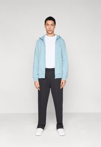 Φούτερ με φερμουάρ σε ανοιχτό γαλάζιο, λευκό μπλουζάκι και σκουρόχρωμο γκρι παντελόνι. Το φούτερ διαθέτει κορδόνια, και η εμφάνιση ολοκληρώνεται με λευκά αθλητικά παπούτσια.