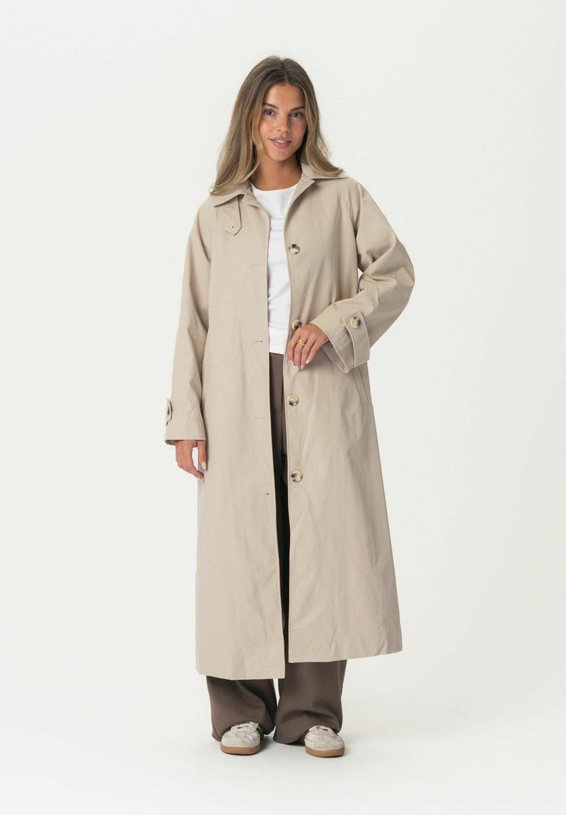 Beige trench coat with a large collar, front buttons, and side pockets. Worn over a white top and brown pants, featuring textured fabric.