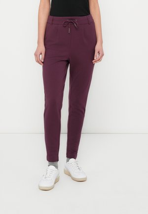 Persoon draagt donkerpaarse slim-fit joggerbroek met trekkoord in de taille, grijze sokken, witte sneakers en een zwart shirt tegen een effen witte achtergrond.