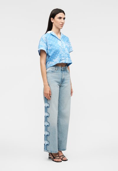 Γυναίκα με μπλε πουκάμισο cropped με σχέδια, ανοιχτό μπλε τζιν παντελόνι καμπάνα με κεντήματα λουλουδιών στα πλάγια και μαύρα σανδάλια με λουράκια, στεκόμενη προφίλ.