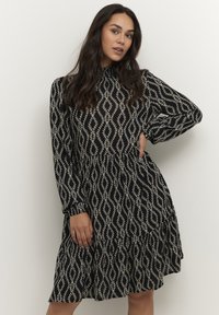 Zwarte jurk met lange mouwen, voorzien van een wit abstract patroon. Het ontwerp heeft een gerimpelde rok en een hoge halslijn. Zachte stoftextuur.