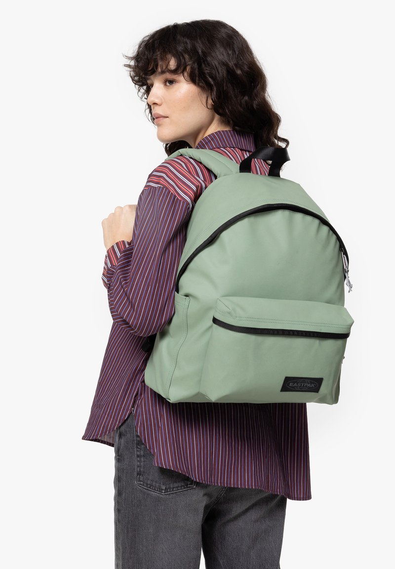 Mochila Eastpak verde con una textura suave, detalles en cremallera negra y un bolsillo frontal. Tiene correas curvadas y una forma redondeada.