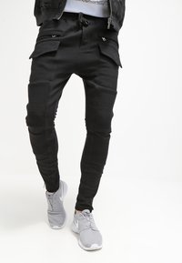 Černé cargo jogger kalhoty s žebrovanými kolenními panely, kapsami na zip a elastickým pasem, v kombinaci se šedými sportovními botami.