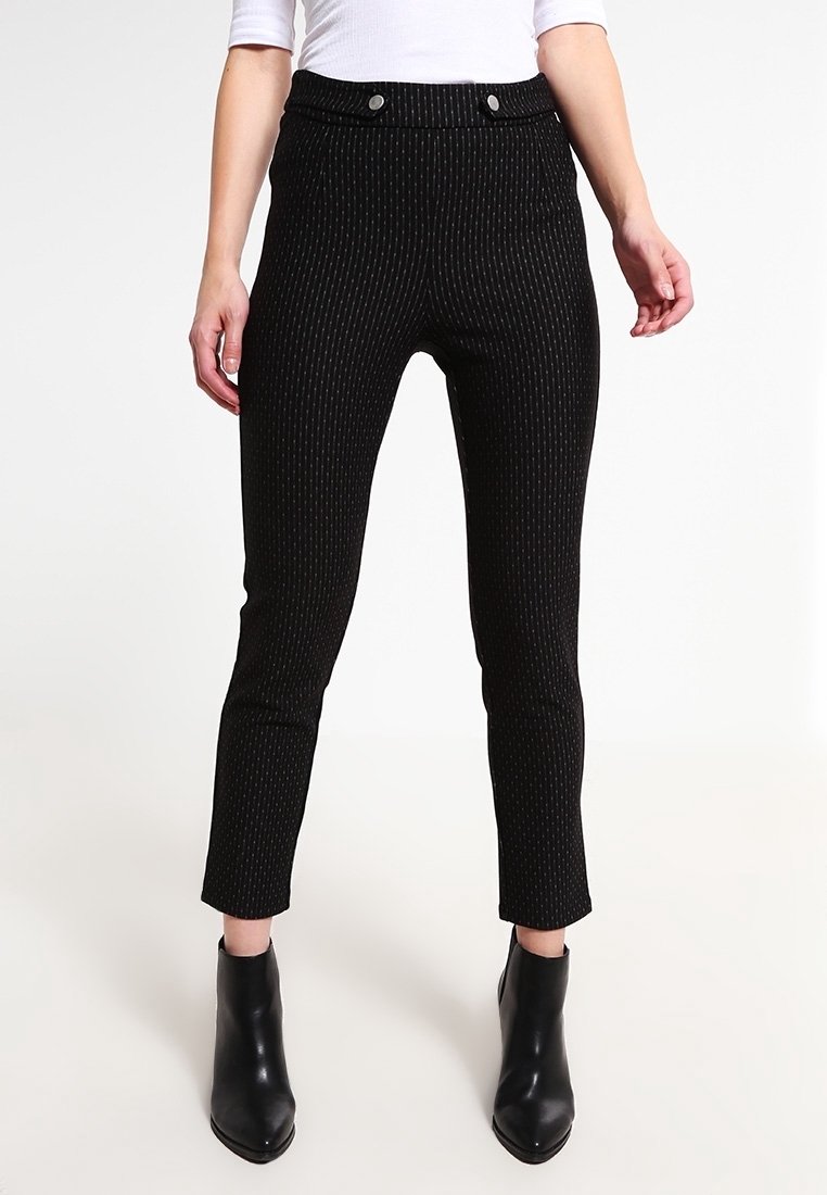 Femme portant un pantalon noir à fines rayures ajusté, longueur cheville, et des bottines noires, debout sur un fond blanc uni.