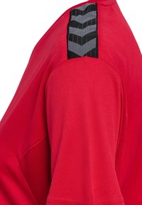 Maglietta sportiva rossa con un accento texturizzato nero sulla spalla con un motivo geometrico. Il tessuto sembra morbido e leggero.