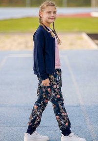 Granatowa kurtka zapinana na zamek, różowa koszulka, dresy w kwiatowy wzór w ciemnoniebieskim kolorze i białe sneakersy. Włosy w warkoczach, stojąca na bieżni.