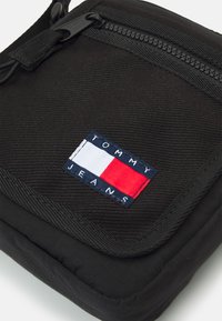 Borsa a tracolla nera in tessuto con superficie texturizzata, chiusura con zip e un'etichetta con blocchi di colore rosso, bianco e blu che recita "TOMMY JEANS."