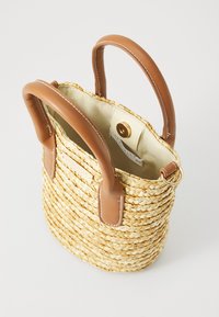Sac en paille tressée avec poignées en cuir marron, forme ronde, doublure beige, fermeture à pression dorée et détail en anneau métallique.
