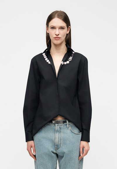 Femme aux cheveux bruns lisses portant une chemise noire à boutons avec un détail de chaîne de perles sur le col et un jean bleu clair avec une ceinture Moschino.
