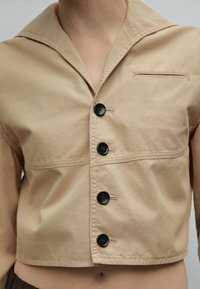 Beige cropped jacket with wide collar and four large black buttons, worn over bare midriff.