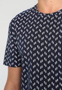 Camisa de color azul marino con mangas cortas, que presenta un patrón de flechas blancas repetido, confeccionada en suave tela de algodón, diseño de cuello redondo.
