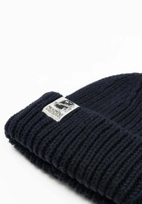 Neulottu laivastonsininen villapipo, jossa on ribbineulos ja käännetty resori; sivussa valkoinen etiketti, jossa on grafiikkaa ja tekstiä.