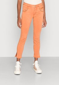 Jean court de couleur pêche avec une texture lisse, poche zippée, et fentes sur le bas, associé à des baskets montantes blanches.