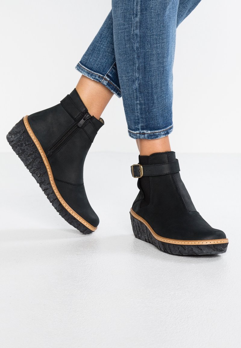 Personne portant des bottines compensées noires avec fermeture éclair latérale et bride à boucle, associées à un jean bleu retroussé sur un sol blanc.
