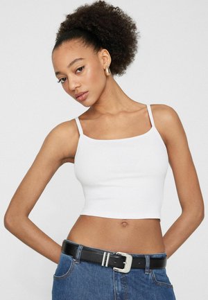 Junge Frau mit lockigem Haar in einem weißen, cropped Tanktop und blauen Jeans mit einem schwarzen Gürtel, die mit den Händen hinter dem Rücken posiert.