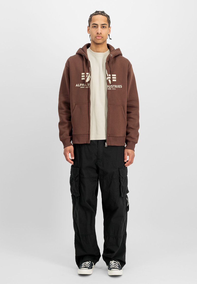 Jeune homme debout portant un sweat à capuche zippé marron Alpha Industries, un t-shirt beige, un pantalon cargo noir et des baskets noires avec des lacets blancs.