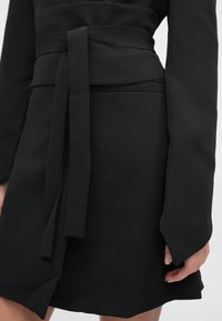 Μαύρο φόρεμα τύπου wrap με μακριά μανίκια, διαθέτει δεμένη ζώνη στη μέση, απαλή υφή και ελαφριά σκίσιμο στο τελείωμα.