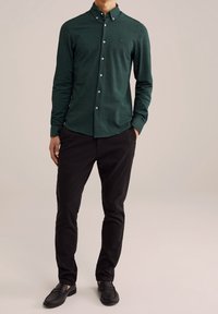 Mörkgrön långärmad skjorta med knäppbar krage, prydd med vita knappar och en strukturerad tyg, parat med svarta byxor och svarta skor.