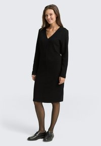 Schwarzes langärmeliges Kleid mit V-Ausschnitt, strukturiertem Stoff, knielangem Schnitt und einer Nahtverarbeitung an der Vorderseite, kombiniert mit schwarzen Schuhen.
