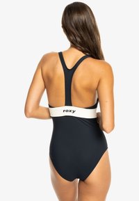 Meriwiktimi navy dengan desain punggung balap dan pita logo putih di pinggang, menampilkan tekstur halus dan siluet yang ramping.