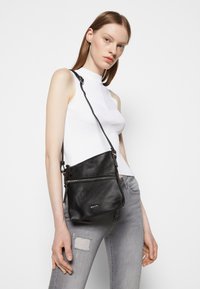 Borsa a tracolla nera in pelle con finitura testurizzata, dotata di chiusura con zip e tracolla regolabile. Modello che la indossa su uno sfondo semplice.