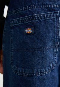 Dark blue denim work pants featuring a back pocket with a rectangular Dickies logo patch. Visible stitching and texture details on fabric.