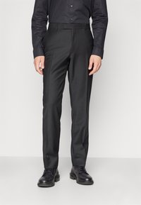 Pantalons noirs ajustés, avec une texture lisse, une taille plate et des passants de ceinture. Portés avec des chaussures à lacets noires.