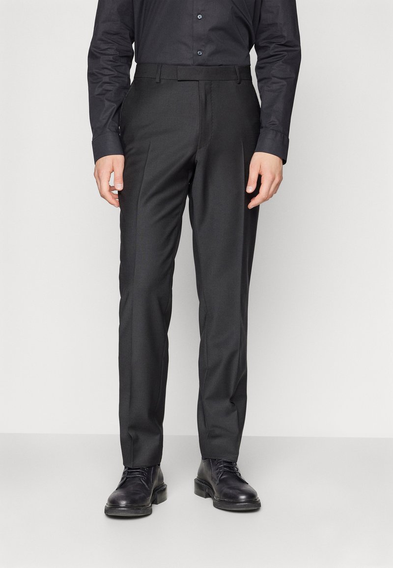 Pantalons noirs ajustés, avec une texture lisse, une taille plate et des passants de ceinture. Portés avec des chaussures à lacets noires.
