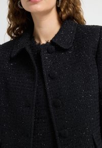 Sort tekstureret jakke med krave, der har et vævet mønster og subtile metalliske tråde. Fem knaplukninger foran, med frynset kanter.