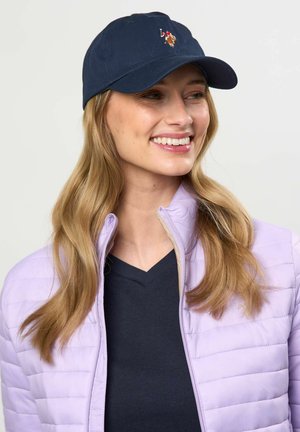 Glimlachende vrouw met lang blond haar, die een marineblauwe pet met logo, een zwart V-hals shirt en een lichtpaarse doorgestikte jas draagt tegen een effen achtergrond.