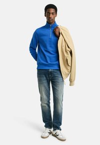 Felpa blu con zip, jeans in denim e sneakers bianche con accenti neri. Giacca beige appoggiata su una spalla. Design semplice e casual.