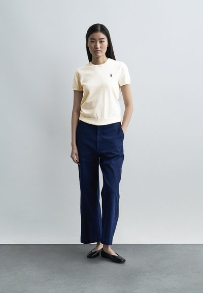 Νέα γυναίκα με μακριά σκούρα μαλλιά φοράει ένα εκρού μπλουζάκι με κοντά μανίκια, μπλε σκούρο φαρδιά παντελόνια και μαύρα παπούτσια μπαλαρίνας μπροστά σε ένα απλό ανοιχτό φόντο.