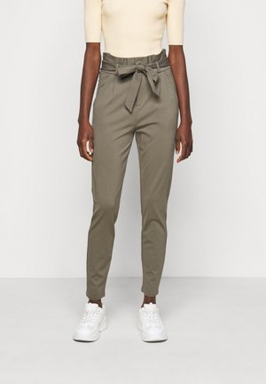 Vrouw draagt beige paperbagbroek met hoge taille en een strikceintuur, gecombineerd met een crèmekleurig T-shirt met korte mouwen en witte sneakers.