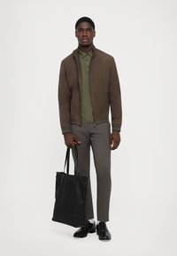 Jachetă bomber din suede maro peste un polo verde, pantaloni gri slim-fit, geantă tote neagră și pantofi negri. Design simplu, aspect casual.