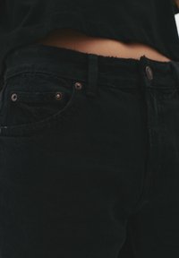 Jean en denim noir avec un ourlet effiloché, des boutons en cuivre et un design classique à cinq poches. Le matériau semble texturé et durable.
