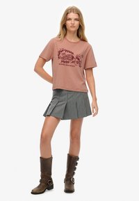Camiseta gráfica color rosa con mangas cortas y un diseño audaz, combinada con una falda gris con rayas y pliegues, y botas marrones hasta la rodilla.