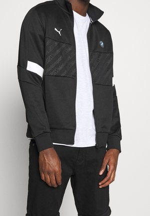 Chaqueta negra con cremallera y acentos blancos, que presenta los logotipos de BMW y Puma, bolsillos texturizados y un acabado de tela suave. Lleva puesta una camiseta blanca.