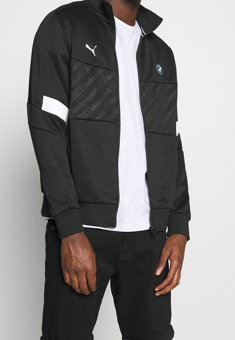 Svart zip-up jacka med vita accenter, prydd med BMW- och Puma-logotyper, texturerade fickor och en slät tygfinish. Bärs över en vit T-shirt.