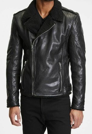Mann trägt eine schwarze Lederjacke mit Lammfellkragen, Reißverschluss vorne, Reißverschlusstaschen und schwarze Hose, gezeigt von Hals bis Hüfte.