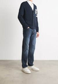 Marinblå stickad cardigan med v-ringning, två framfickor, vit bokstaverad emblemlogga, i kombination med blå jeans och vita sneakers.