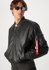 Black leather bomber jacket with ribbed collar and cuffs. Features a zippered pocket on the sleeve and a red "REMOVE BEFORE FLIGHT" tag.